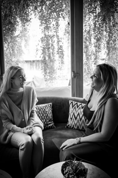 Zwei Frauen sitzen auf einer Couch und sprechen miteinander in einem lichtdurchfluteten Raum.